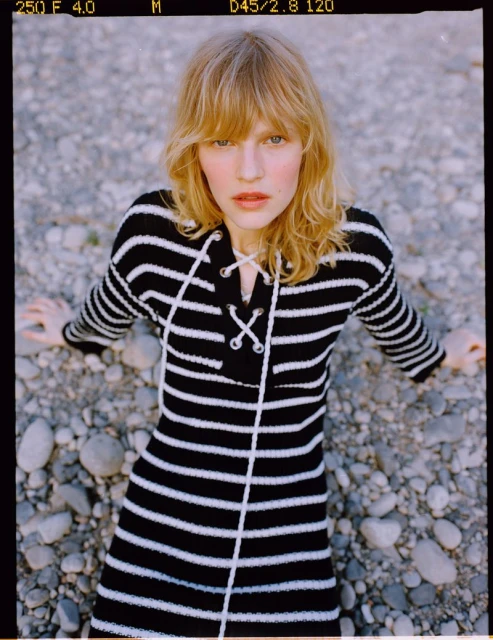 Blonde woman in a striped sweater dress poses outdoors on a rocky surface, looking directly at the camera with a neutral expression.