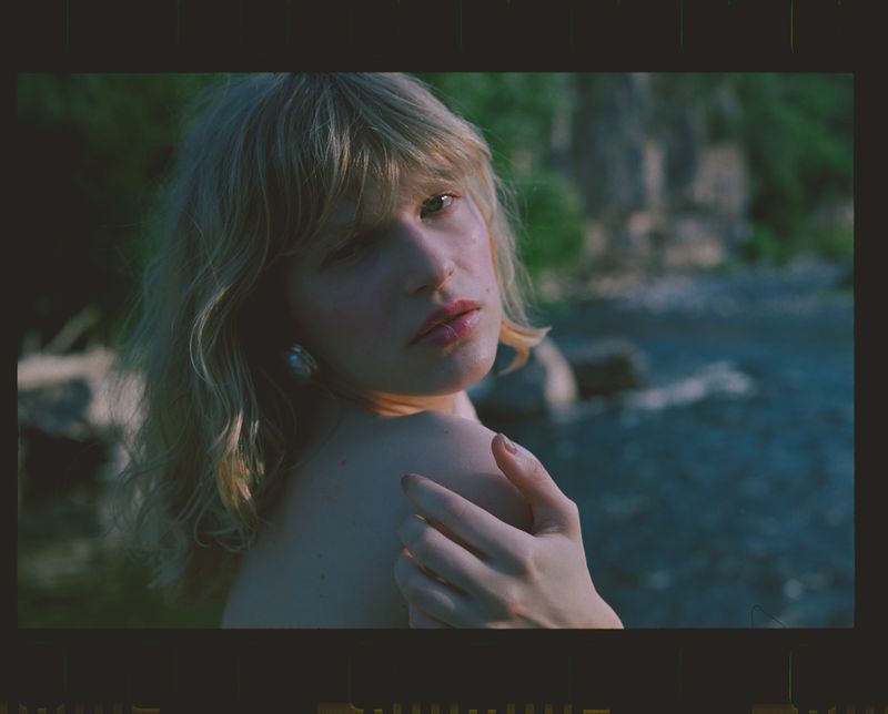 Portrait of a young adult woman with blonde hair and bare shoulders, gazing towards the camera against a natural background of water and greenery.