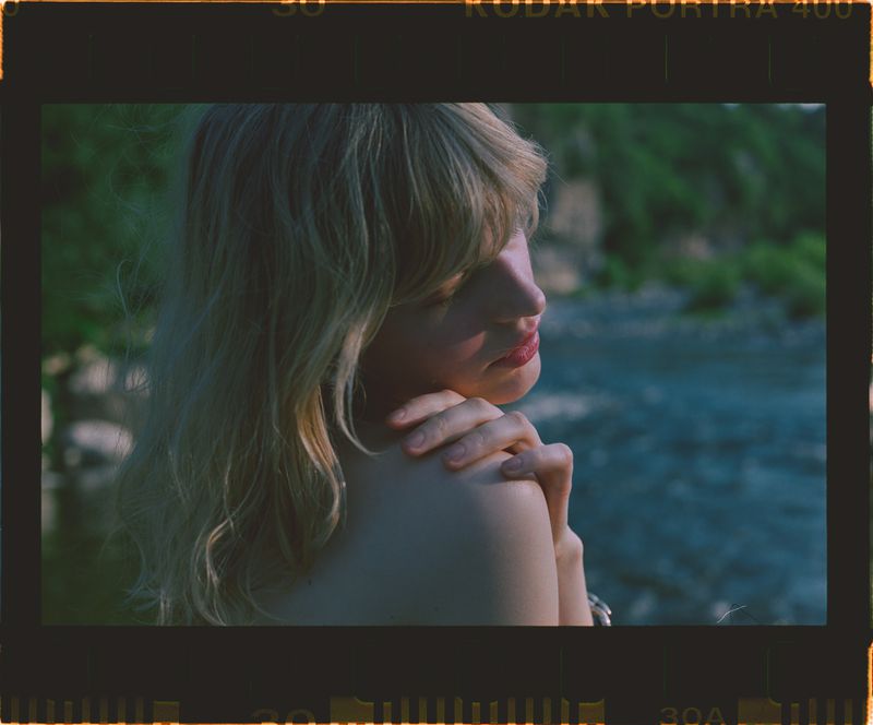 A blonde woman with red lipstick poses against a river backdrop, showcasing shoulder and arm, with lush green background and film border.