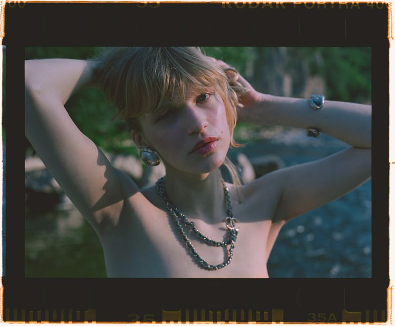 Young woman with bare shoulders, raises her hands behind her head, wearing jewelry, standing in a natural outdoor setting. 