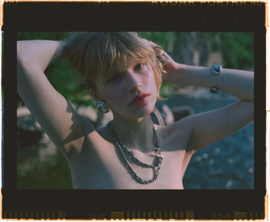 Young woman with bare shoulders, raises her hands behind her head, wearing jewelry, standing in a natural outdoor setting. 
