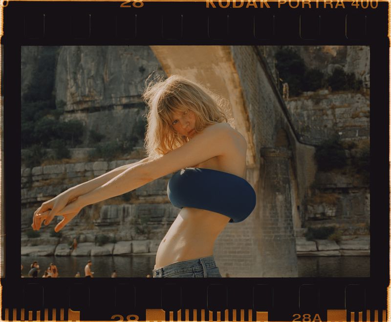 Young woman in blue top poses outdoors with rock cliffs and water. Sunlit scene with film photography aesthetic.