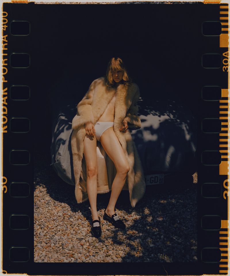 Blonde woman wearing coat and underwear sitting next to a white car in outdoor setting. Dark brown frame with Kodak Portra 400.