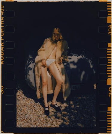 Blonde woman wearing coat and underwear sitting next to a white car in outdoor setting. Dark brown frame with Kodak Portra 400.