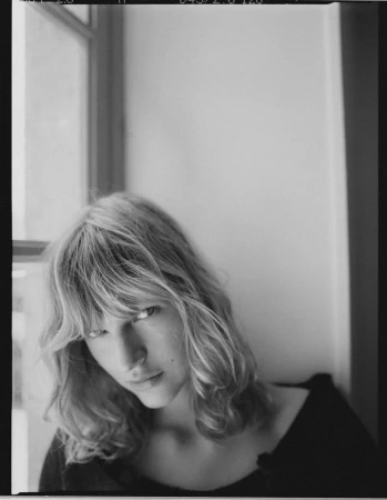 Portrait of a blonde woman with bangs looking down towards the camera against a neutral indoor wall with a window.