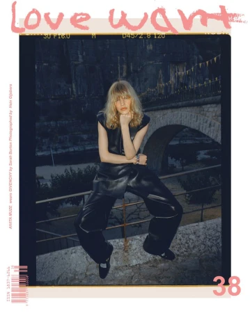Editorial fashion shot of a model wearing a dark outfit, posing outdoors against an old stone bridge and wall in a cool, muted palette.