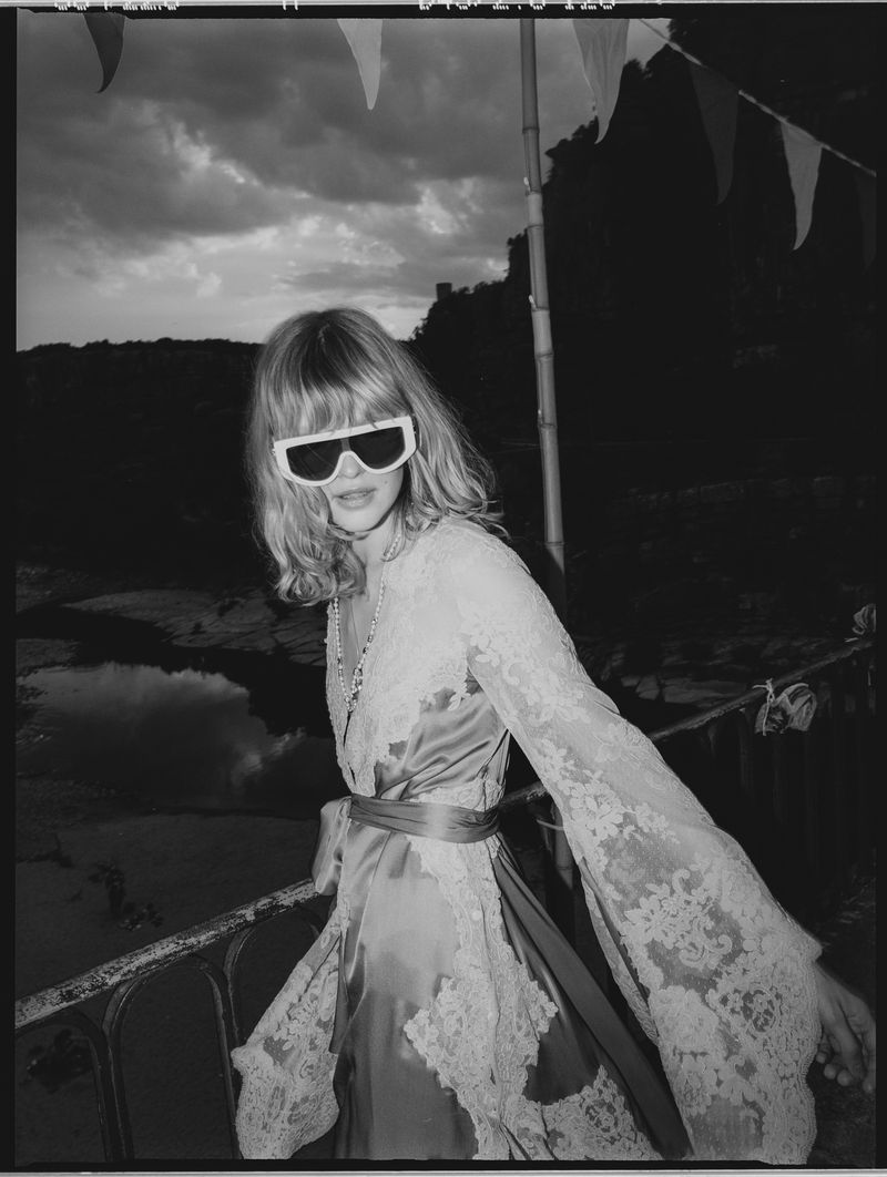 A woman with blond hair stands near water wearing a lace dress and sunglasses in a black and white portrait on an overcast day.
