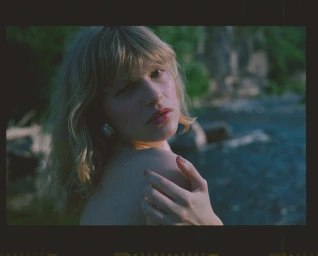Close-up portrait of a woman with blonde hair looking over her shoulder outdoors near water.