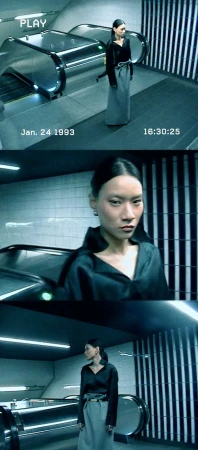 A woman stands near an escalator in an underground station setting, wearing a long skirt and collared shirt with a blurred, vintage effect.