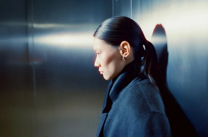 Profile view of a woman with dark hair pulled back, wearing a dark coat, standing against a metallic wall, illuminated by a bright light.