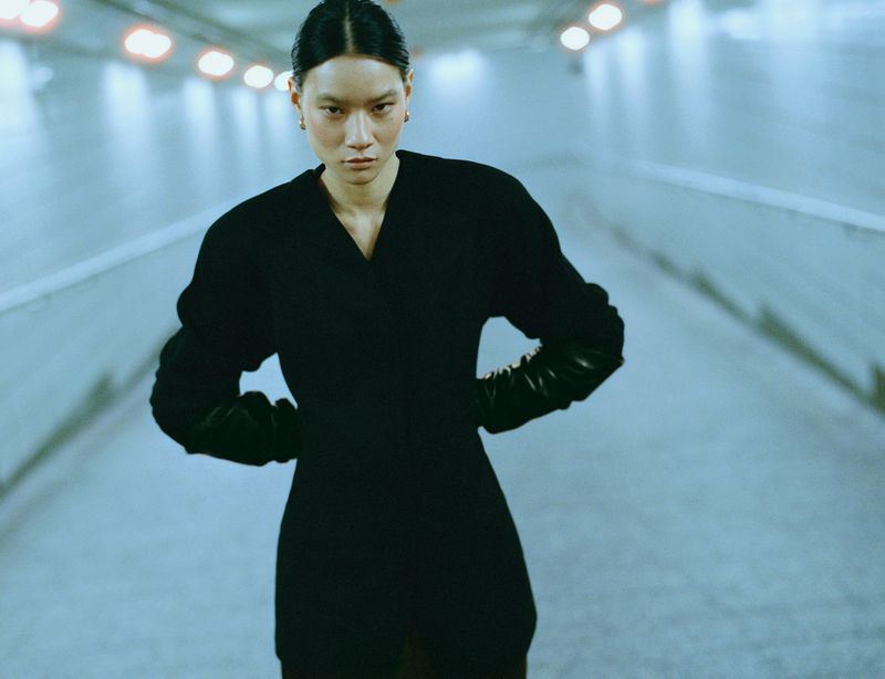 Asian woman with hands on hips poses in a black dress and gloves, illuminated by overhead lights in a concrete tunnel.