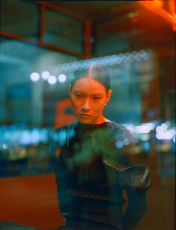 Portrait of an Asian woman with slicked-back hair, wearing a dark top, reflected in a glass window with blurred lights in the background.
