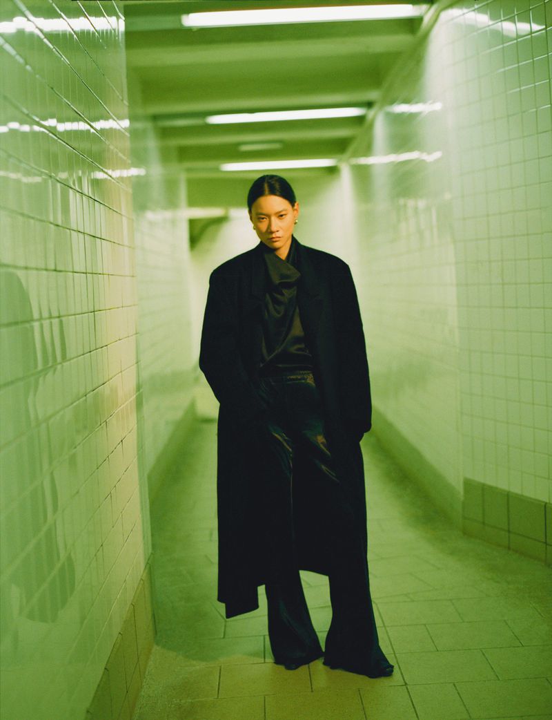 An Asian woman in a long black coat and black pants poses in a green-tiled pedestrian tunnel with fluorescent lighting.