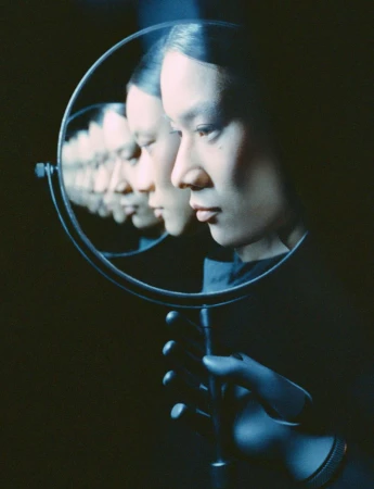 Multiple reflections of an adult woman's face are captured in a circular mirror held by a gloved hand against a dark background.