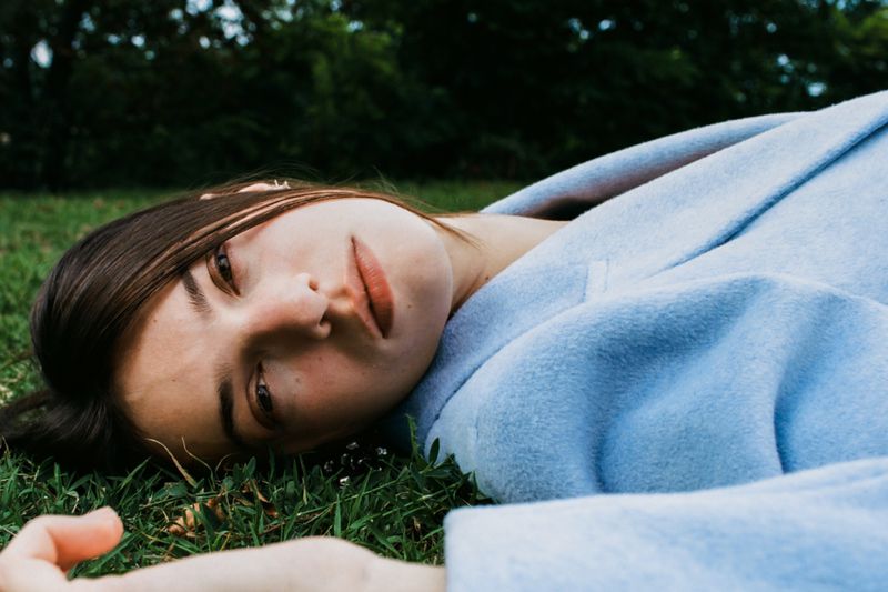 Young woman with dark hair in a light blue coat lies on green grass, looking directly at the camera in a relaxed, outdoor setting.
