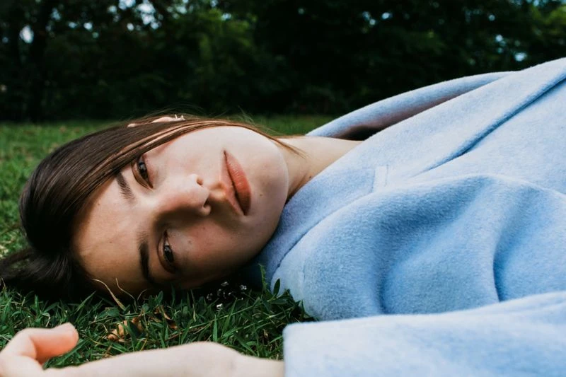 Young woman with dark hair in a light blue coat lies on green grass, looking directly at the camera in a relaxed, outdoor setting.