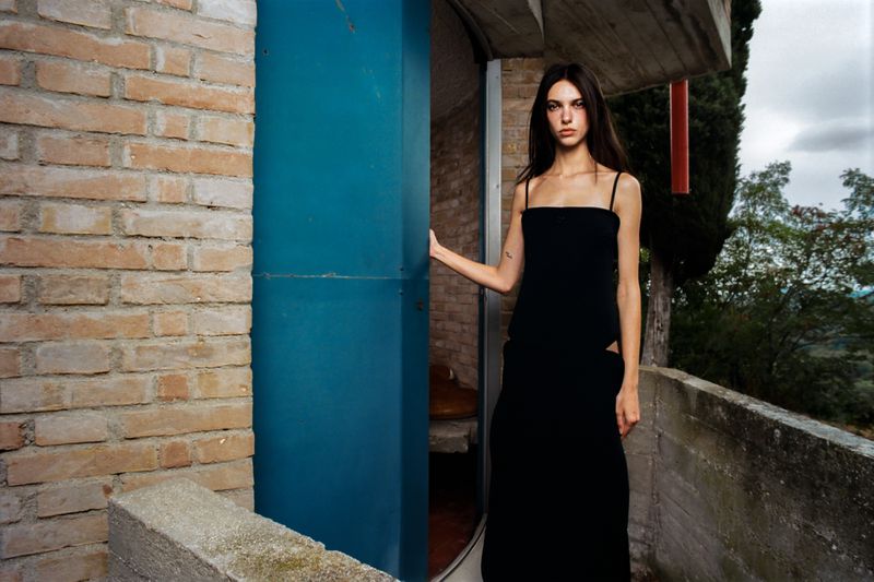An adult female fashion model wearing a black dress stands by a blue doorway with a brick wall, set against an outdoor greenery background.