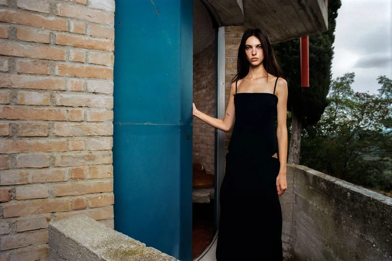 An adult female fashion model wearing a black dress stands by a blue doorway with a brick wall, set against an outdoor greenery background.