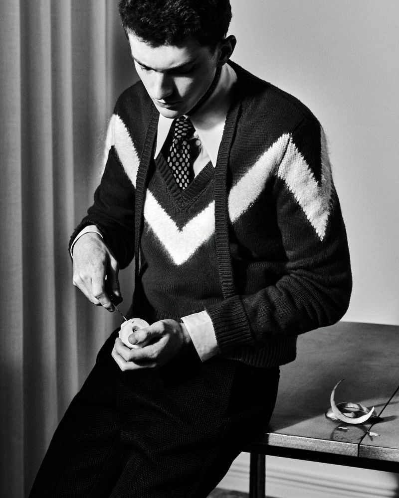 Monochrome portrait of a young man in a patterned sweater peeling a fruit at a table in an indoor setting with neutral backgrounds.