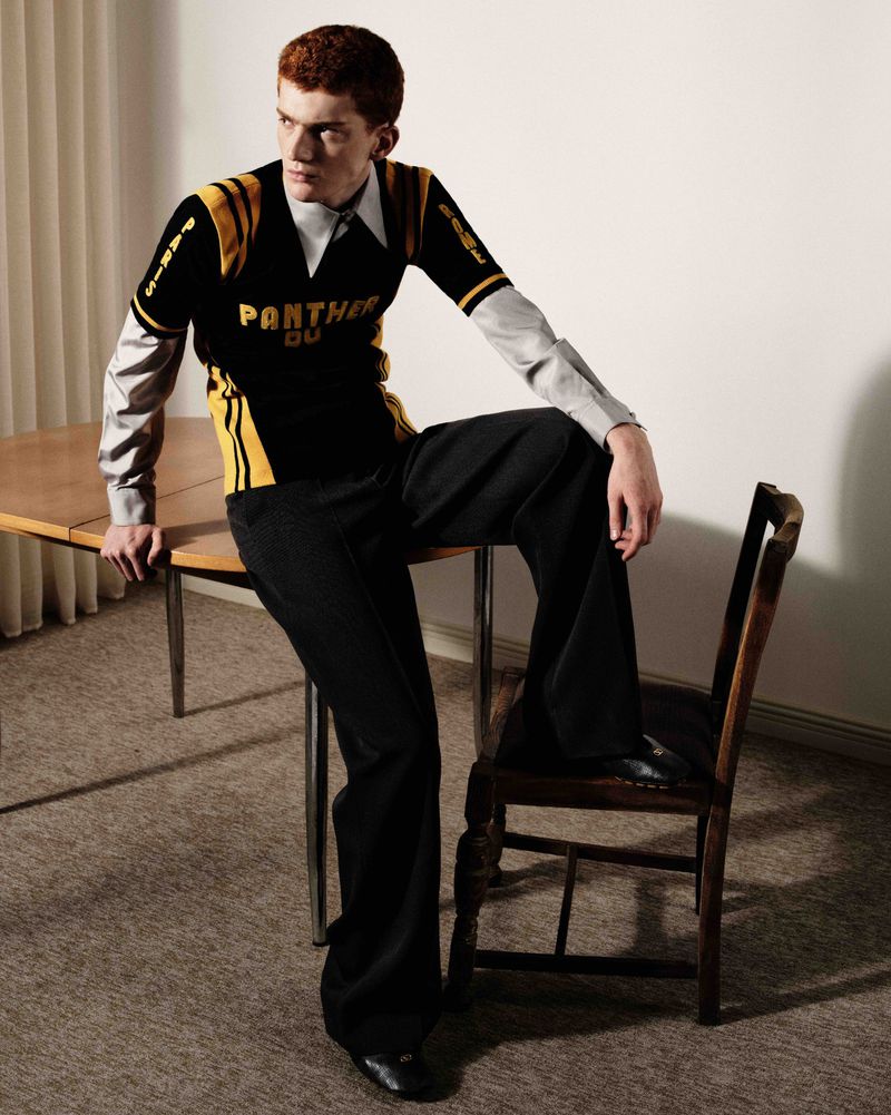 Stylish man in a black and gold jersey and wide-leg pants poses indoors, seated on a vintage chair with a table beside him.