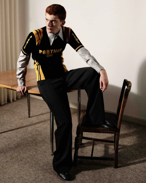 Stylish man in a black and gold jersey and wide-leg pants poses indoors, seated on a vintage chair with a table beside him.