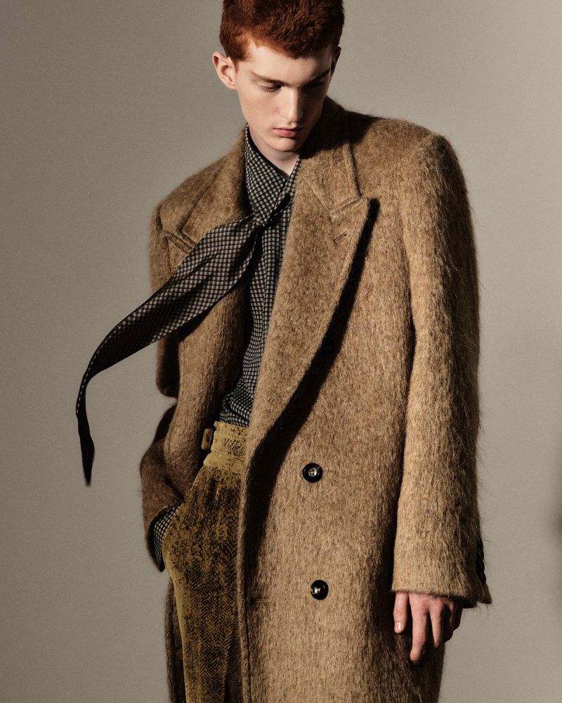 Fashion portrait of a red-haired male model wearing a brown wool coat, patterned shirt, and necktie against a neutral backdrop.