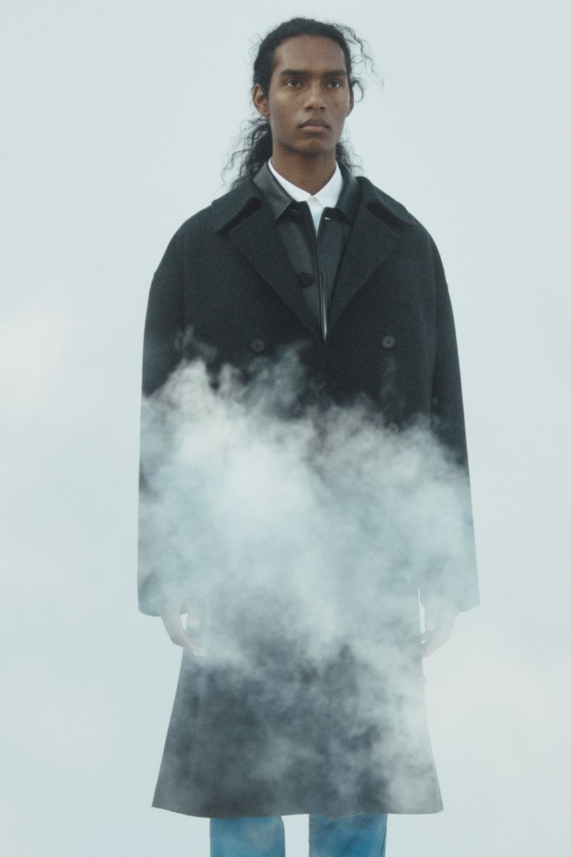 Young man in a dark overcoat stands in a foggy, atmospheric setting, creating a striking and enigmatic fashion portrait.