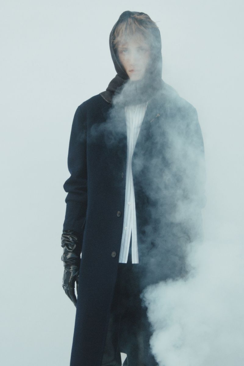 A single person in a dark hooded coat stands amidst smoke, creating a dramatic and mysterious fashion-forward editorial portrait.
