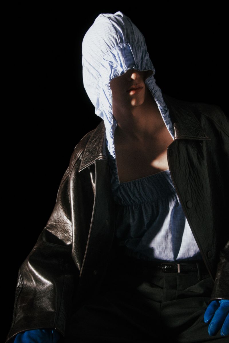 Model in dramatic lighting wearing a leather jacket and draped blue fabric over their head, set against a dark, stark background.