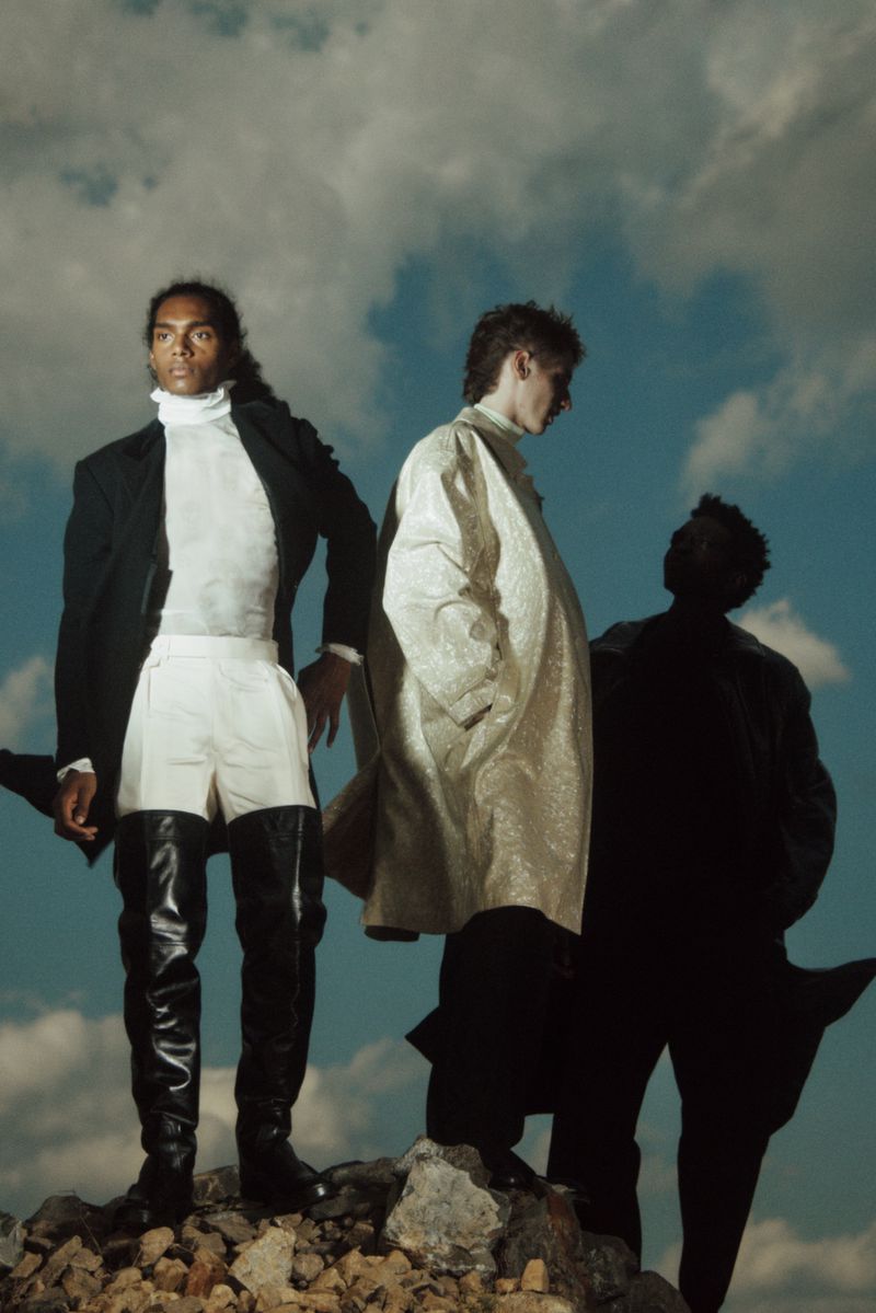 Three fashion models strike a pose outdoors, showcasing black and gold jackets, white pants, and over-the-knee boots against a partly cloudy sky.