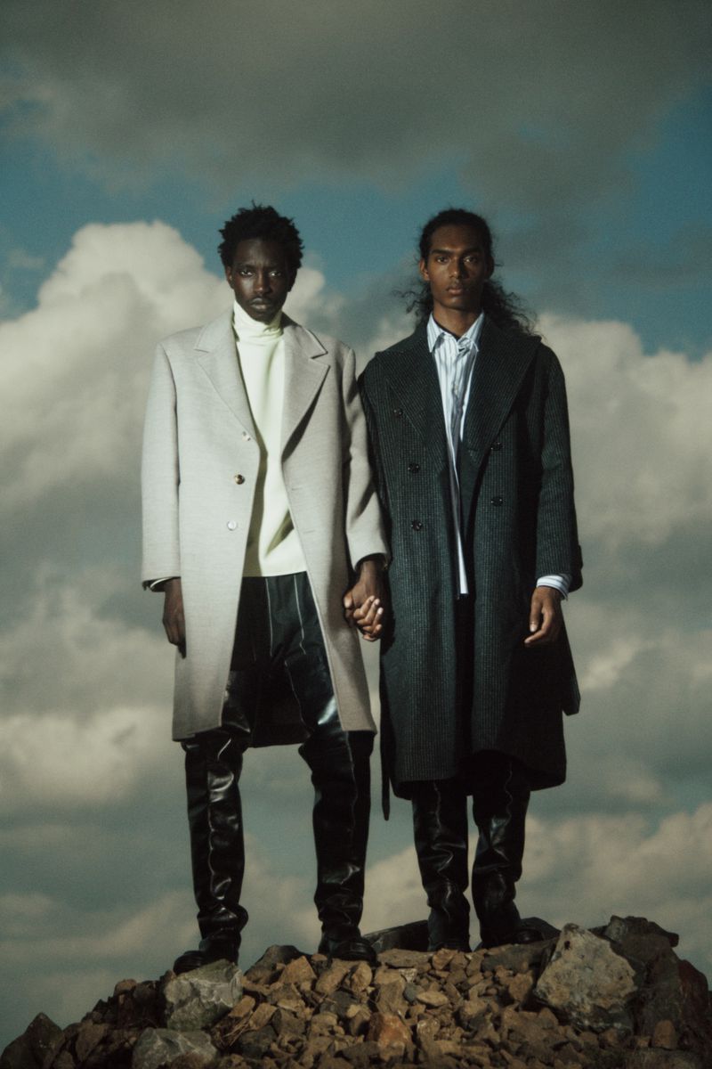 Two Black male fashion models pose on rocks wearing long coats, turtleneck, and shiny pants against a cloudy sky in an outdoor editorial shot.