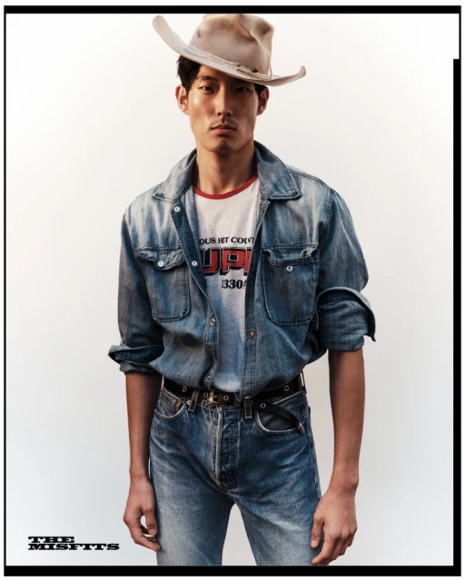 Portrait of Asian male model in denim shirt and cowboy hat against plain background in a fashion editorial shot.