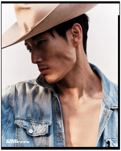 East Asian male model wearing a tan cowboy hat and an open light-wash denim shirt against a white backdrop.