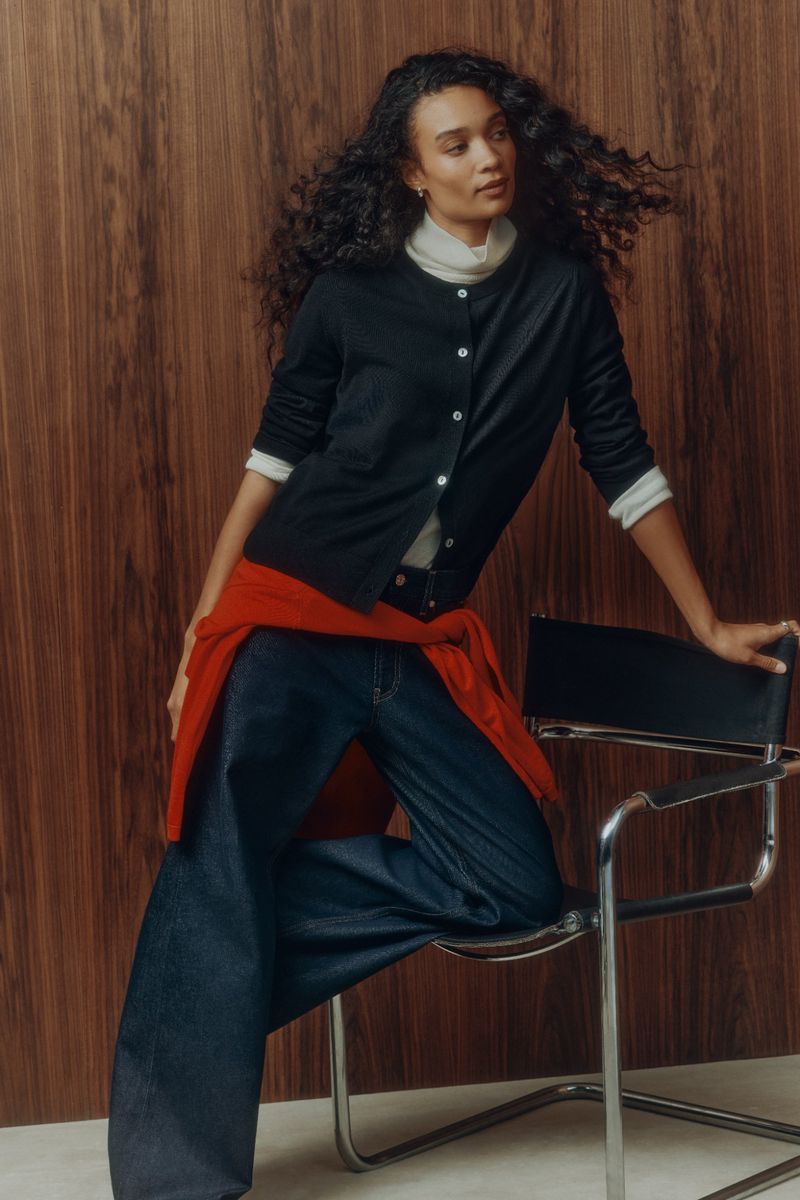 Woman with long dark curly hair wearing a dark cardigan, white turtleneck, and blue jeans, posing near a chrome chair.