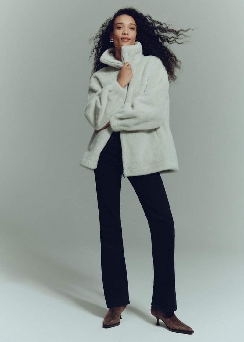 Portrait of a woman in an ivory jacket, black pants, and heels, posing in a photo studio with a neutral backdrop, fashion editorial.