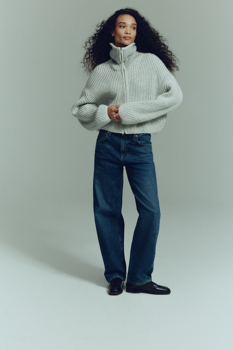 A woman with dark curly hair poses in a pale green zip-up sweater, dark blue jeans, and black shoes against a grey backdrop.