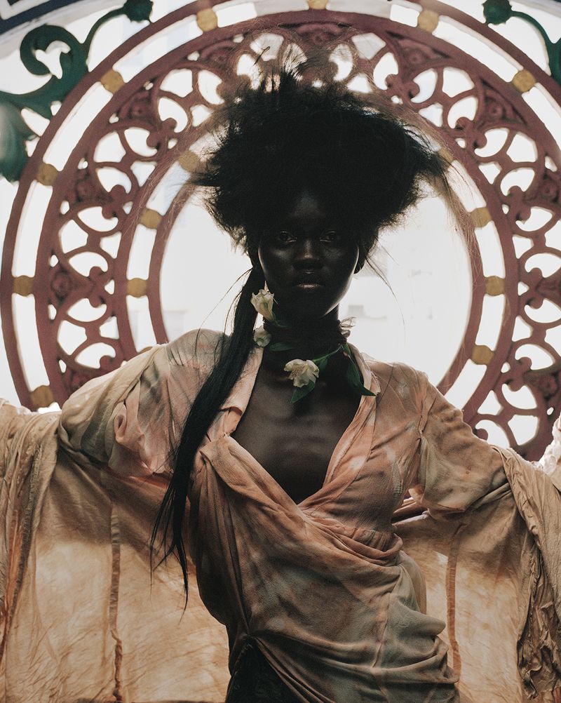An adult African woman in a draped peach dress poses in front of an ornate window, showcasing fashion and beauty in an indoor setting.
