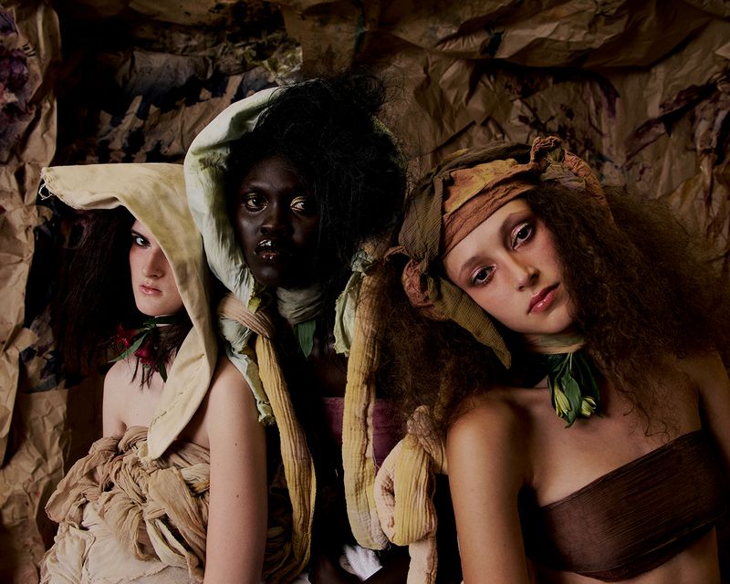 Three women pose in conceptual fashion. Dark hair, tan and dark skin tones, earth-toned fabrics, textured backdrop, shallow depth-of-field.