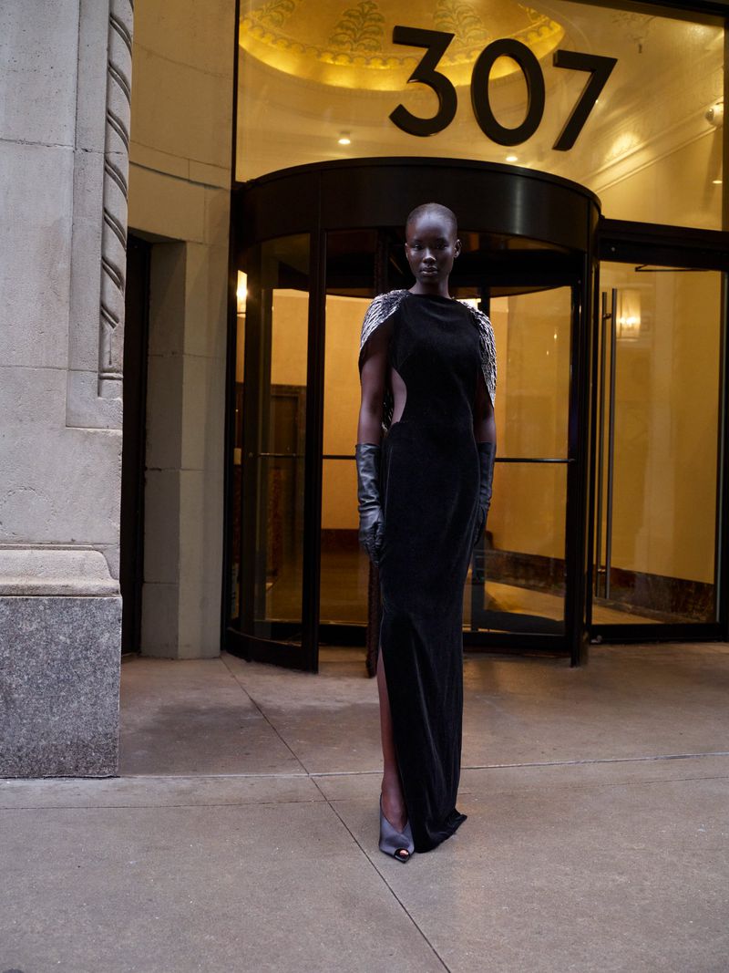 Elegant black woman in long black gown poses outside building with revolving doors and '307' sign, accessorized with gloves.