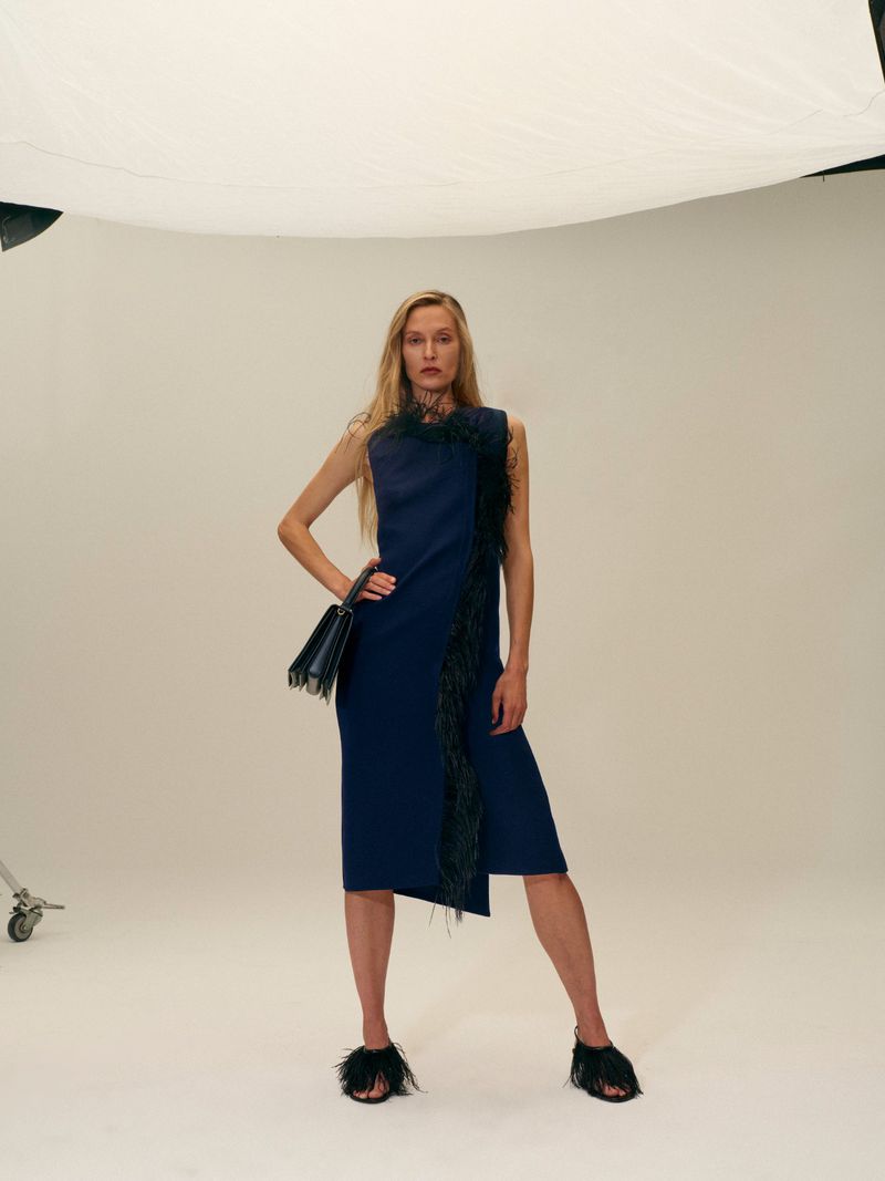 A fair-skinned female fashion model poses in a sleeveless navy midi dress with black feather trim and sandals, holding a black handbag in a studio.