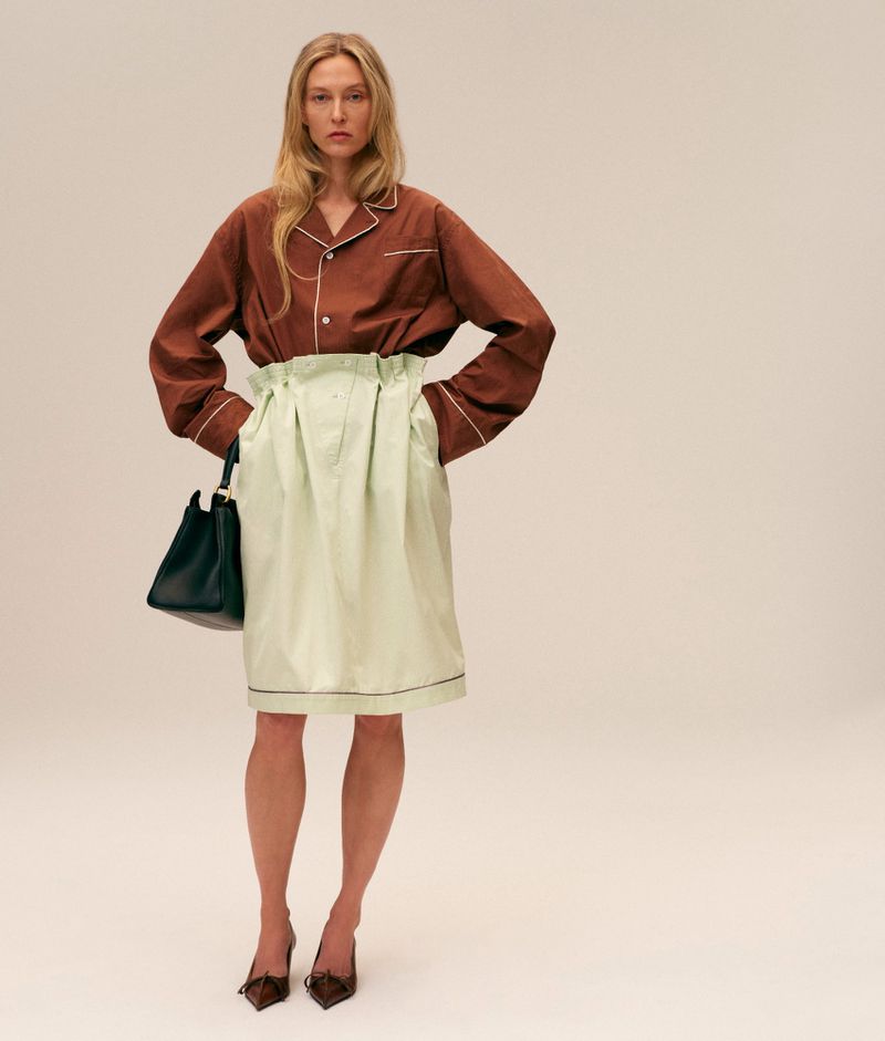 A blonde adult woman is posing in a brown top, light-green skirt, and brown heels, holding a dark-green handbag in a studio setting.