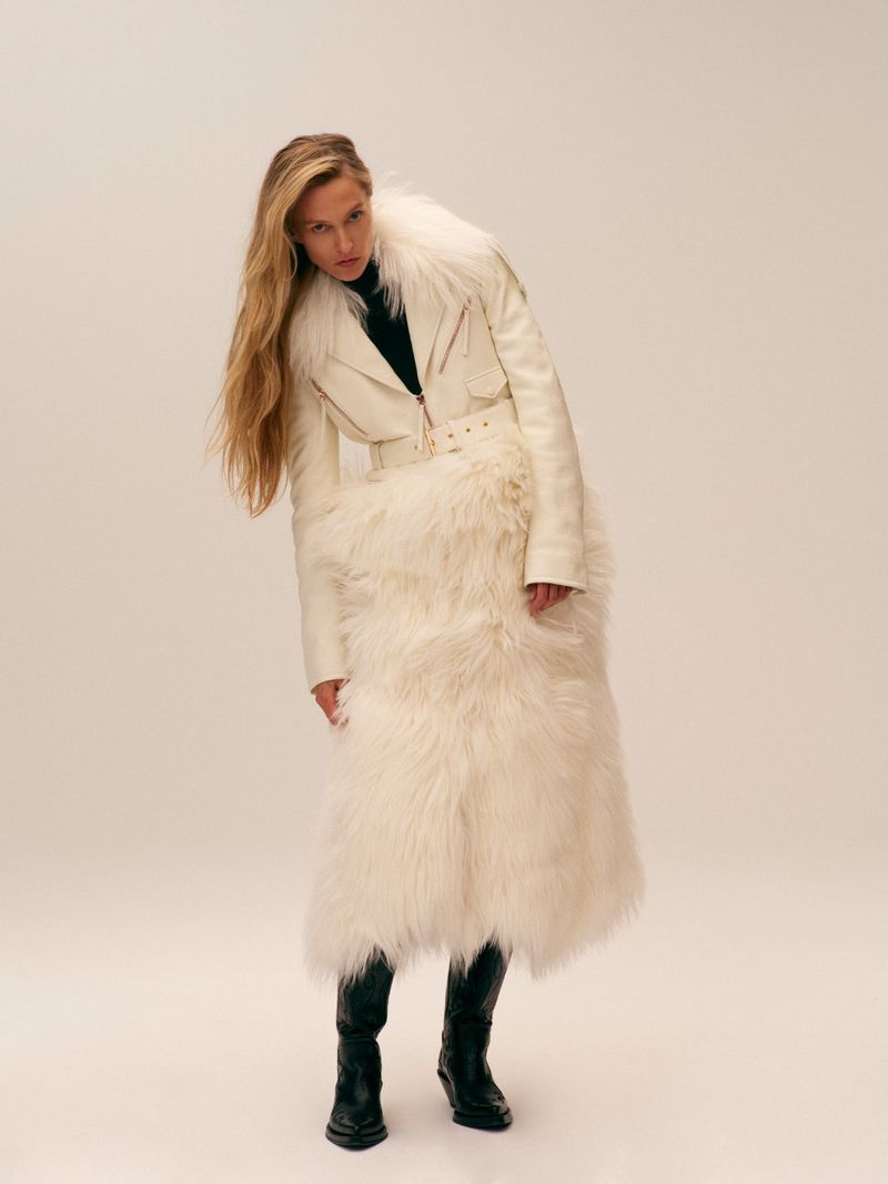 A woman poses indoors wearing a white jacket with fur details, a black top, and black boots against a neutral backdrop.