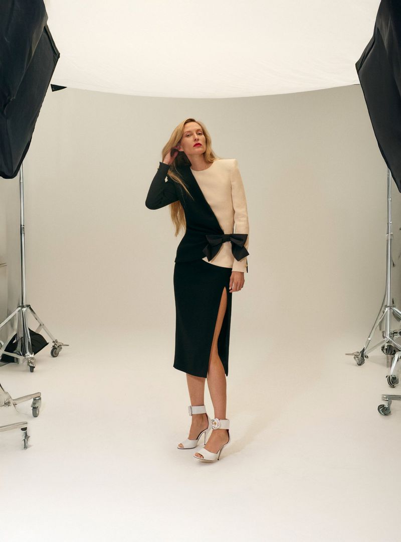 A full shot of a woman in a fashionable cream-and-black dress with a bow, complemented by white high heels, posing in a photo studio.