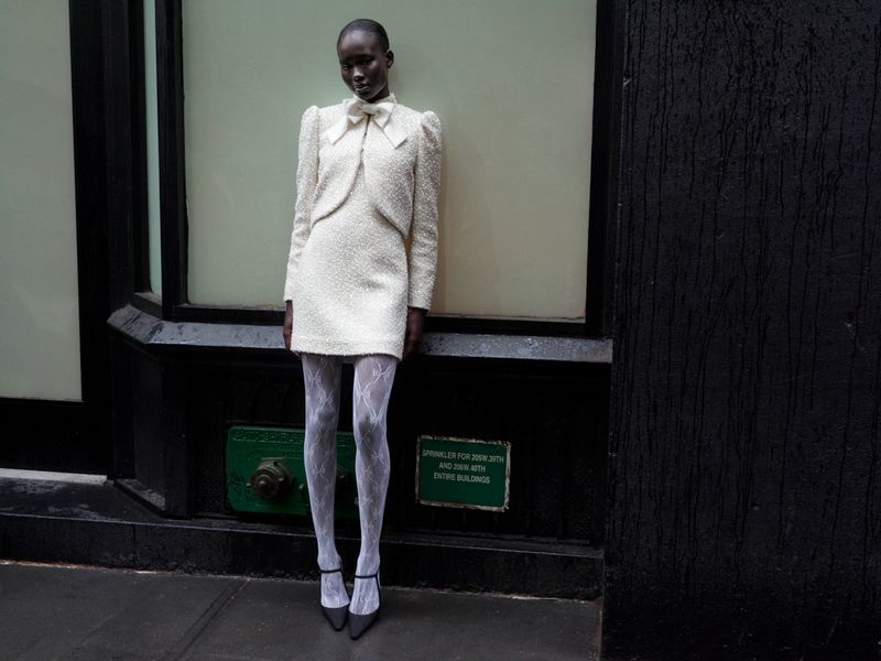 A slender Black woman poses in a cream tweed mini dress, sheer tights, and heels against a grey city wall with black trim and accents.