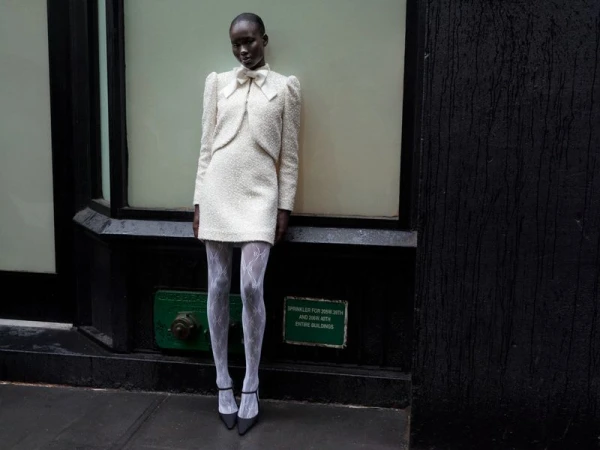 A slender Black woman poses in a cream tweed mini dress, sheer tights, and heels against a grey city wall with black trim and accents.