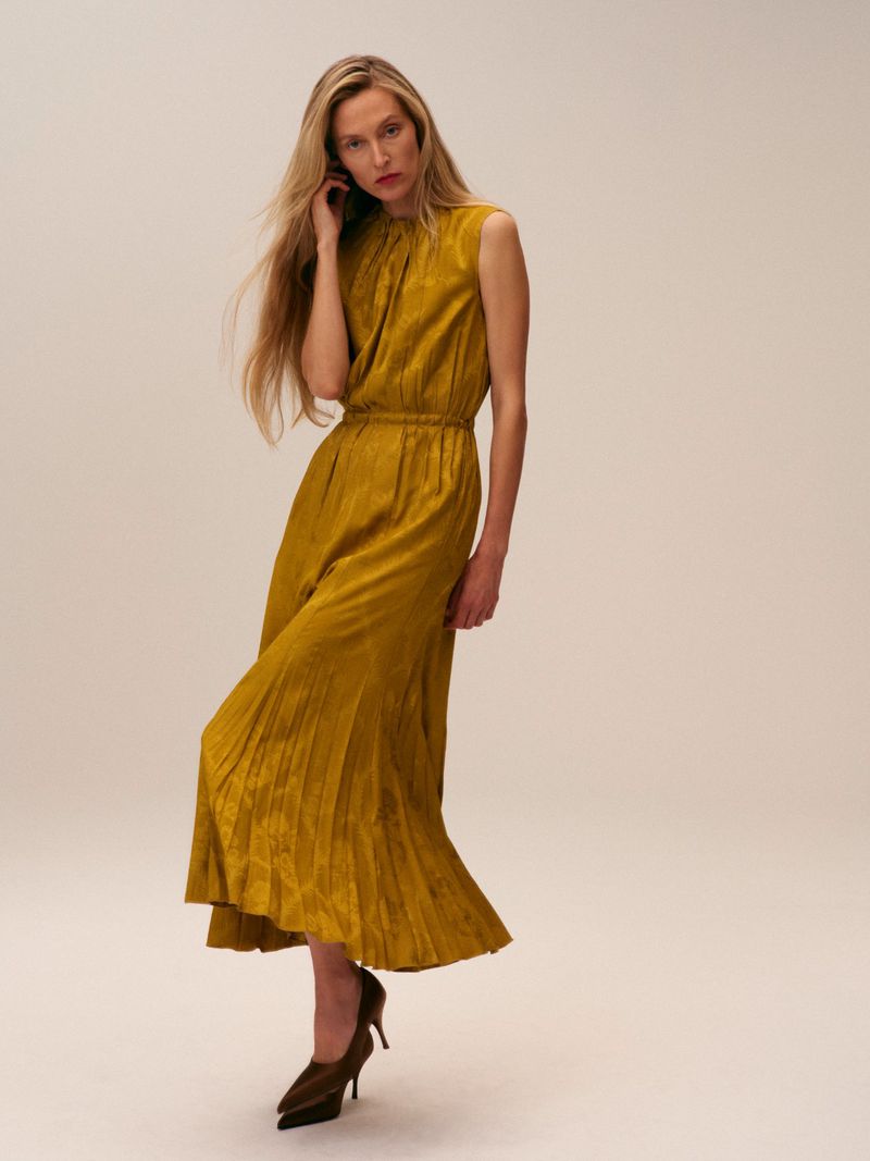 A slender woman poses in a textured yellow sleeveless dress and brown heels against a light background in a studio shot.