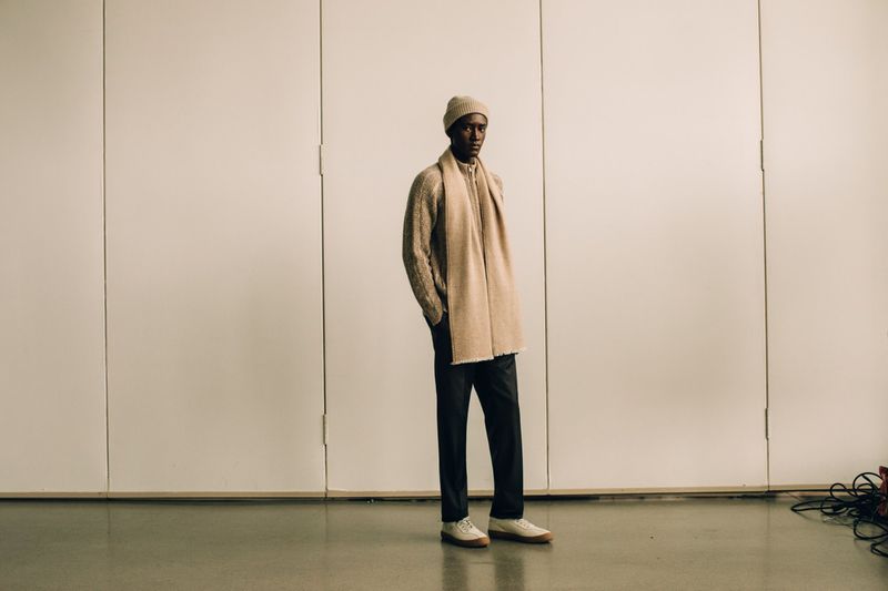 A full shot of an adult male model in a beige knit beanie, scarf, and sweater, dark pants, and sneakers standing in front of a white wall.