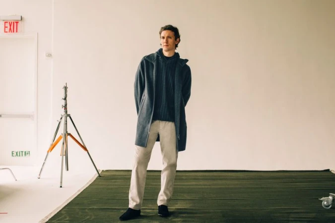 A man in a gray coat and sweater stands in a photo studio against a white backdrop with a green carpet and photographic lighting.