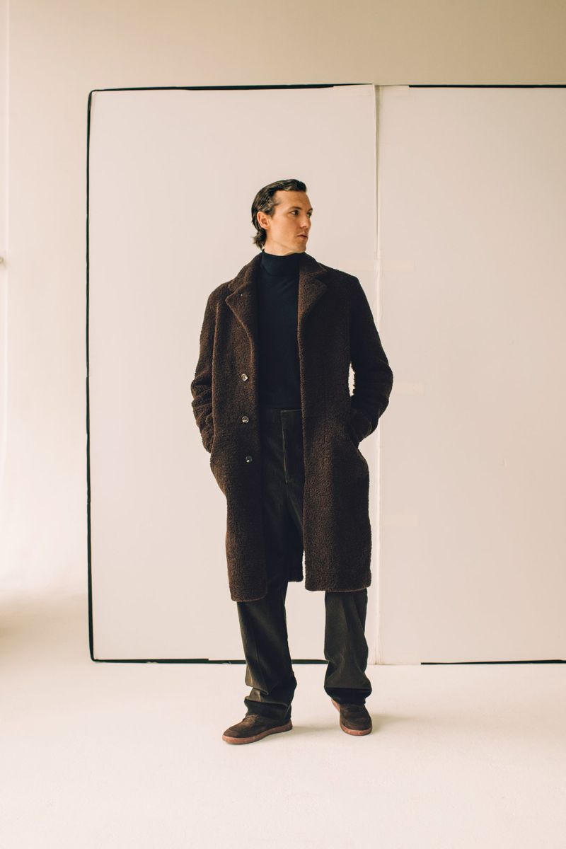 An adult male model poses in a dark brown coat, black turtleneck, and dark pants in a full-body shot against a white backdrop in a studio.