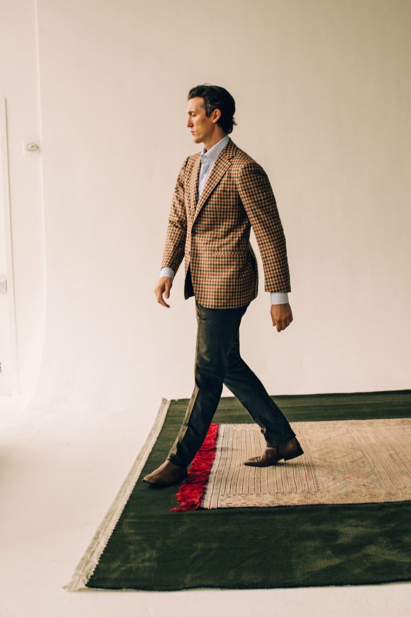 A stylish man in a patterned blazer walks across a modern rug in a brightly lit studio, showcasing men's fashion and interior design.